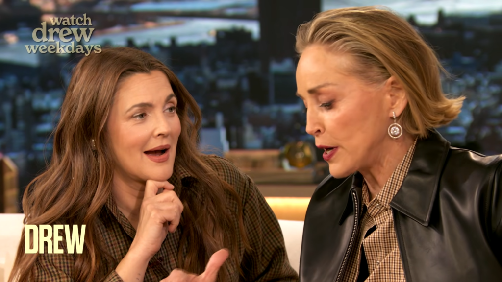 Two women sit close together on a talk-show set, mid-conversation; left is a brunette in plaid, right is a blonde with a leather jacket and statement earrings.