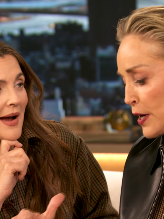 Two women sit close together on a talk-show set, mid-conversation; left is a brunette in plaid, right is a blonde with a leather jacket and statement earrings.