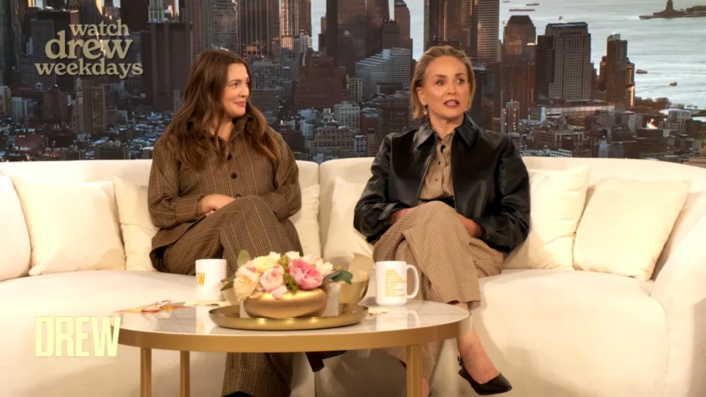 Two women sit on a cream couch during a talk-show interview set with a city skyline backdrop and daisies on a table in front of them, speaking softly to camera alternating between chairs.