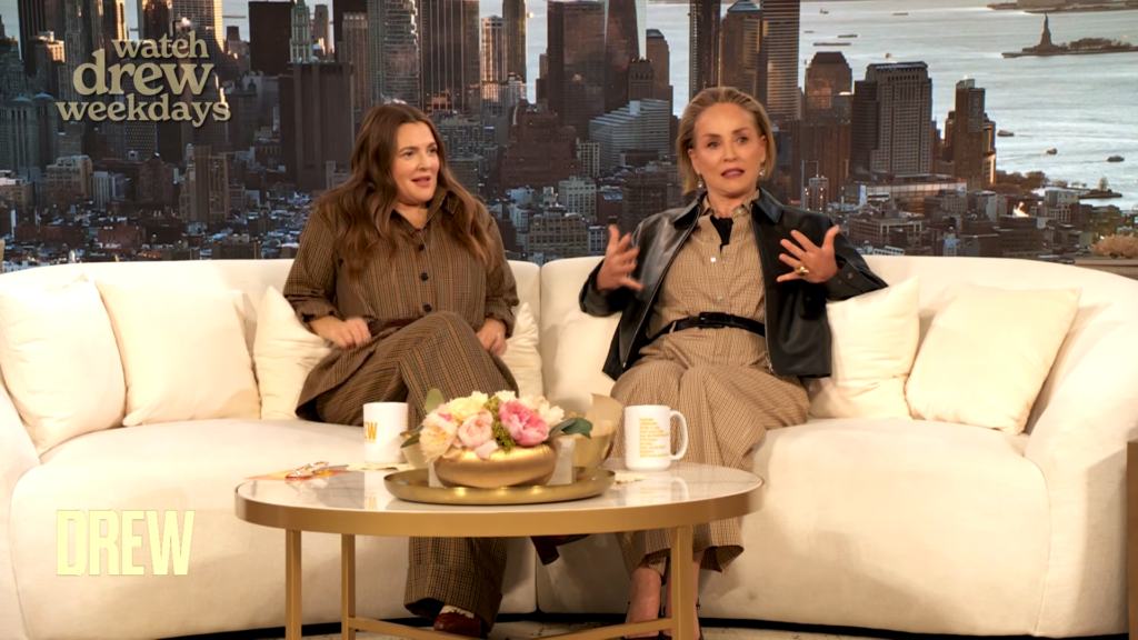 Two women sit on a cream sofa on a talk-show set, with a city skyline backdrop and coffee mugs in front of them on a glass table.