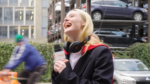 Young woman laughing during an on-street interview; headset around her neck and a microphone visible in the foreground.
