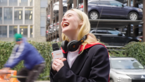Young woman laughing during an on-street interview; headset around her neck and a microphone visible in the foreground.