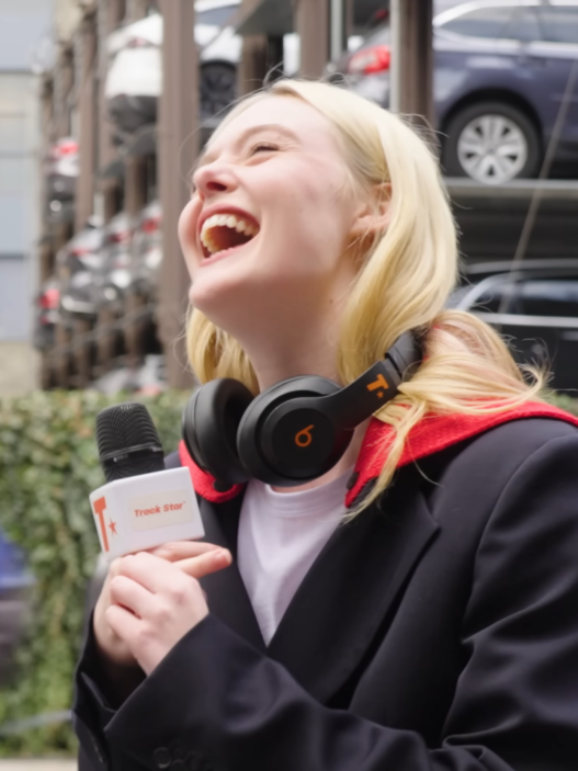 Young woman laughing during an on-street interview; headset around her neck and a microphone visible in the foreground.