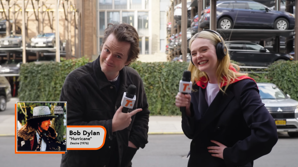 Two reporters interview on a city street, holding mics; a Bob Dylan 'Hurricane' promo card is in the foreground.