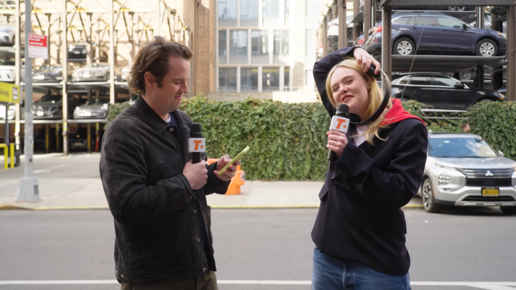 Two reporters outdoors on a city street, holding branded microphones; man checks his phone while a woman speaks, with parked cars and a parking structure in the background.