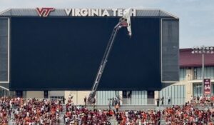 Crane reaches the top of a Virginia Tech stadium scoreboard as workers adjust the VT sign while a crowd fills the stands below.
