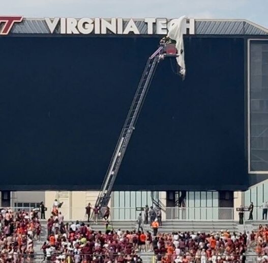 Crane reaches the top of a Virginia Tech stadium scoreboard as workers adjust the VT sign while a crowd fills the stands below.