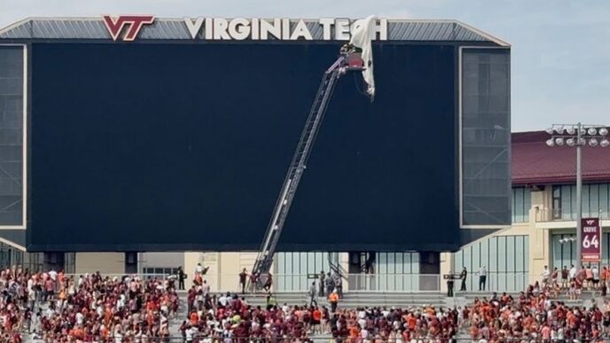 Crane reaches the top of a Virginia Tech stadium scoreboard as workers adjust the VT sign while a crowd fills the stands below.