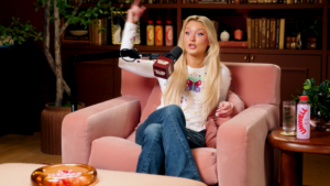 Blonde woman in a white sweater speaks into a desk microphone on a podcast set, seated on a pink chair with bookshelves behind her.