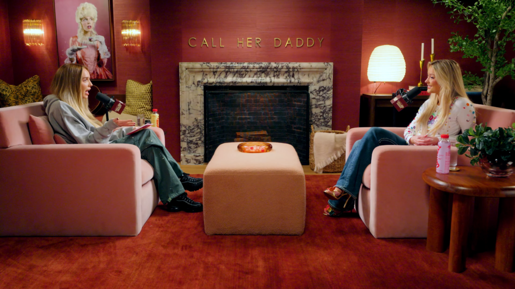 Two women sit on pink chairs facing each other, talking into microphones in a pink lounge studio with a marble fireplace and a 'CALL HER DADDY' sign above it.