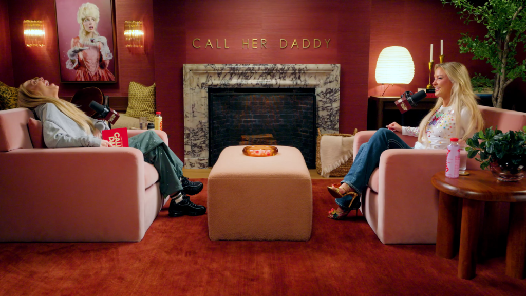 Two women sit on pink chairs in a podcast studio, microphones in front of them, with a 'CALL HER DADDY' sign above a marble fireplace behind them.