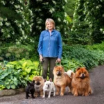 Smiling woman in a denim shirt standing with six dogs in a lush garden.