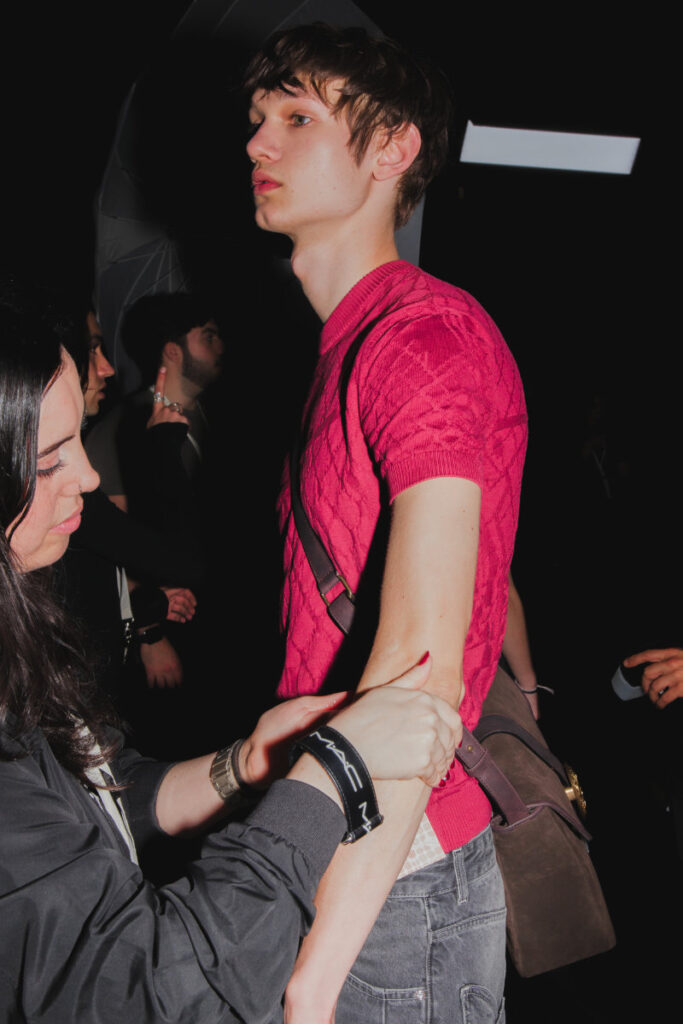 Backstage scene: a model in a bright pink knit shirt stands while a stylist adjusts his sleeve nearby.
