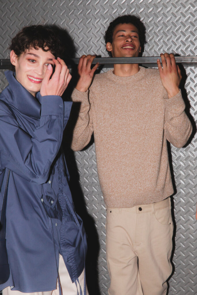 Two friends posing and smiling in front of a textured metal diamond-plate wall; the man on the right holds a bar across his shoulders.