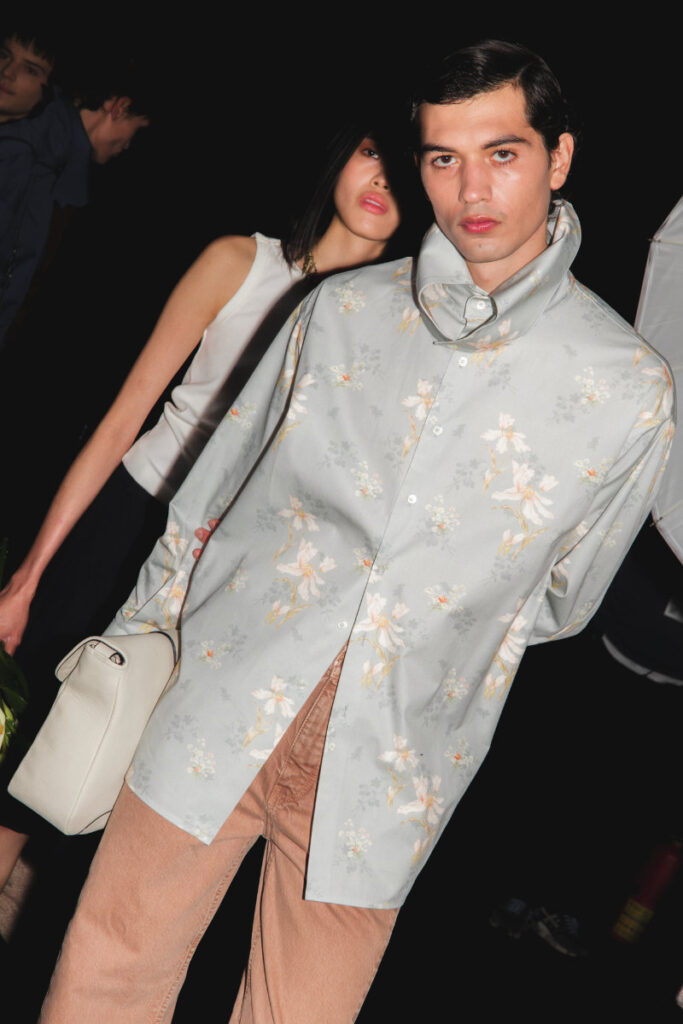 Male model in a pale blue floral shirt and tan pants, holding a white bag, posing at a night fashion event.