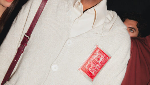 Male model in a cream cardigan with a red stamp-like patch, white shirt underneath, maroon shoulder strap visible; dark background with a female model partially seen on the left.