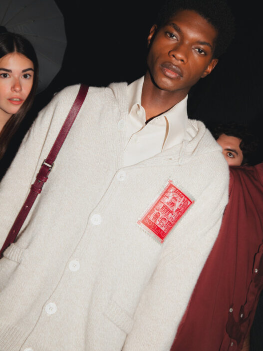 Male model in a cream cardigan with a red stamp-like patch, white shirt underneath, maroon shoulder strap visible; dark background with a female model partially seen on the left.