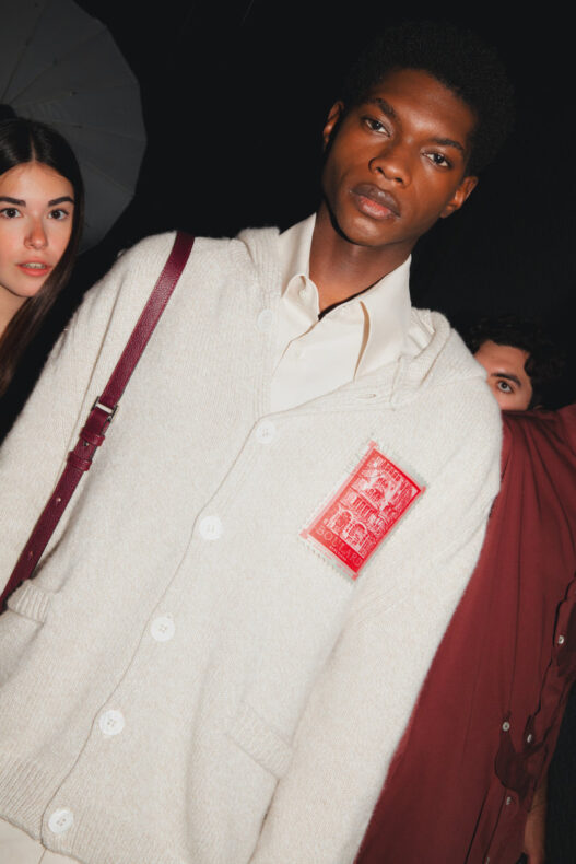 Male model in a cream cardigan with a red stamp-like patch, white shirt underneath, maroon shoulder strap visible; dark background with a female model partially seen on the left.