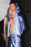 Model in metallic blue satin hooded jacket and matching pants, unbuttoned to reveal the chest and a brown pendant necklace (backstage).