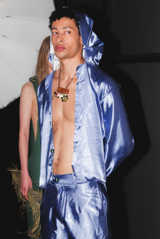 Model in metallic blue satin hooded jacket and matching pants, unbuttoned to reveal the chest and a brown pendant necklace (backstage).