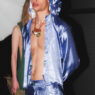 Model in metallic blue satin hooded jacket and matching pants, unbuttoned to reveal the chest and a brown pendant necklace (backstage).