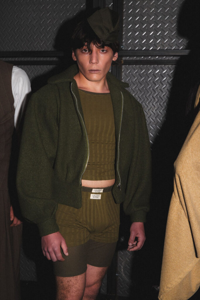 Person wearing an olive-green cropped top, high-waisted shorts, and an oversized zip-up jacket with a cap, standing against a metal diamond-plate wall.