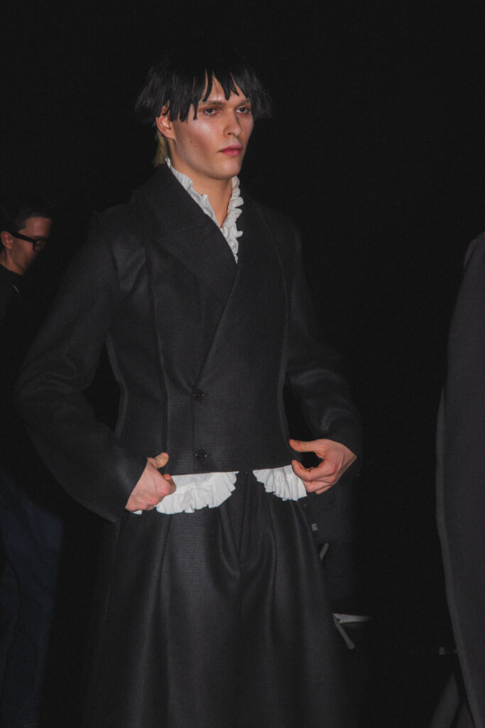 Model in a black Victorian-inspired coat with a white ruffled shirt, on a runway.