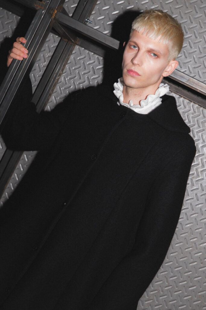 Fashion model with short platinum hair wearing a black coat and white frilled collar, leaning against a metal diamond-plate wall.