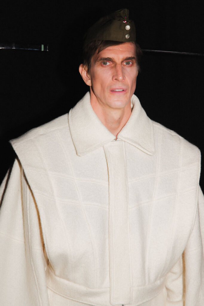 Model wearing a heavy cream wool cape-like coat and a small military-style hat at a fashion show.