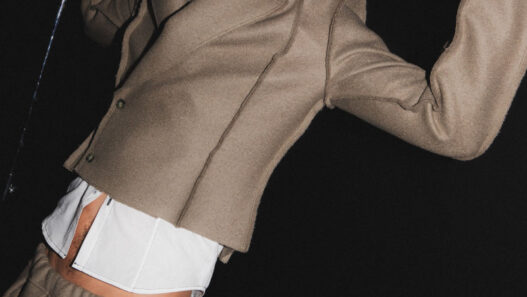 Male fashion model in a beige tailored suit with a white shirt, posed dramatically against a dark backdrop, holding a metallic rod above his head.