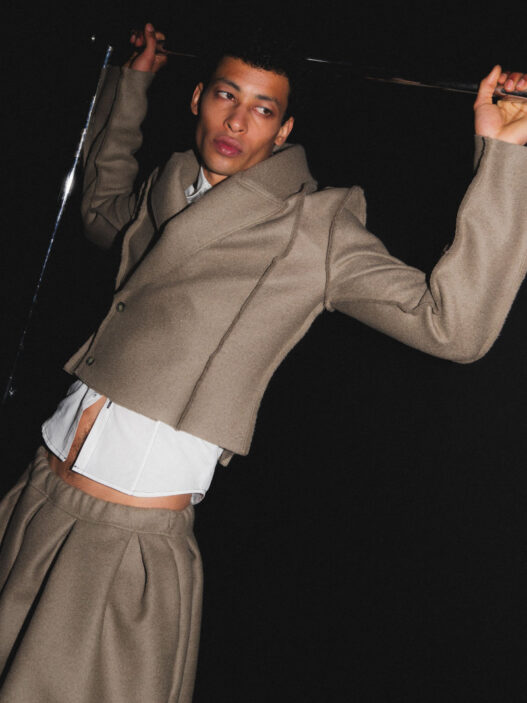 Male fashion model in a beige tailored suit with a white shirt, posed dramatically against a dark backdrop, holding a metallic rod above his head.