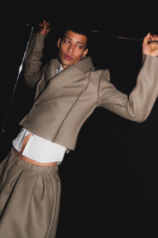 Male fashion model in a beige tailored suit with a white shirt, posed dramatically against a dark backdrop, holding a metallic rod above his head.