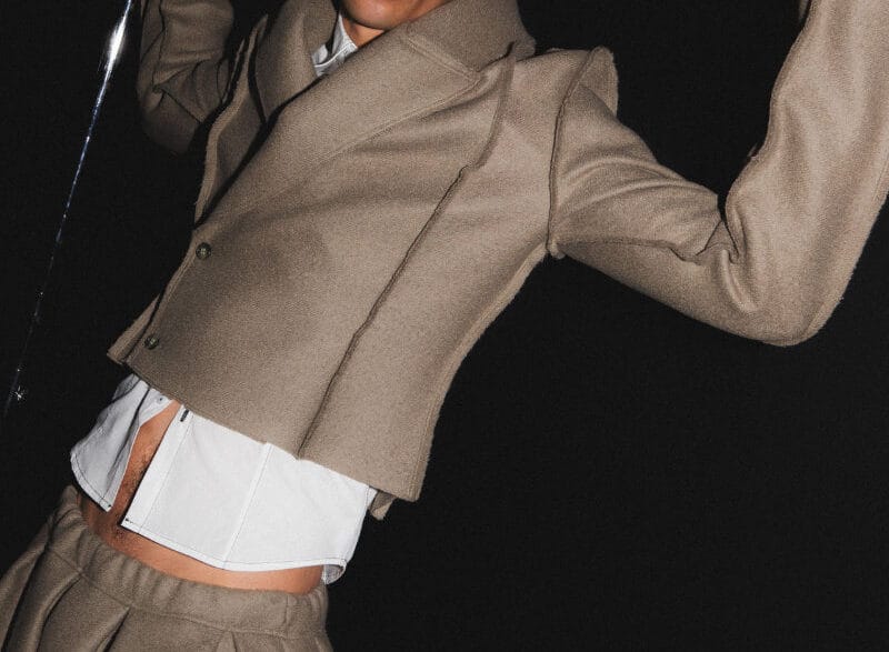 Male fashion model in a beige tailored suit with a white shirt, posed dramatically against a dark backdrop, holding a metallic rod above his head.