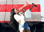 Female performer on stage wearing a white blouse and ripped denim shorts, shouting into a bright red megaphone while holding a microphone.