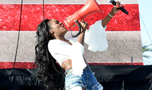 Female performer on stage wearing a white blouse and ripped denim shorts, shouting into a bright red megaphone while holding a microphone.