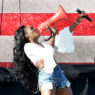 Female performer on stage wearing a white blouse and ripped denim shorts, shouting into a bright red megaphone while holding a microphone.