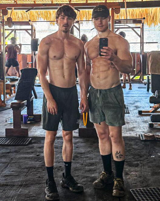 Two shirtless men posing for a mirror selfie in a gym, with others working out in the background and gym equipment around them.