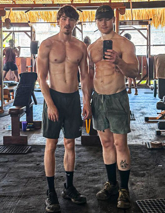 Two shirtless men posing for a mirror selfie in a gym, with others working out in the background and gym equipment around them.