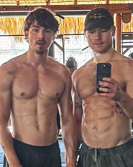 Two shirtless men posing for a mirror selfie in a gym, with others working out in the background and gym equipment around them.