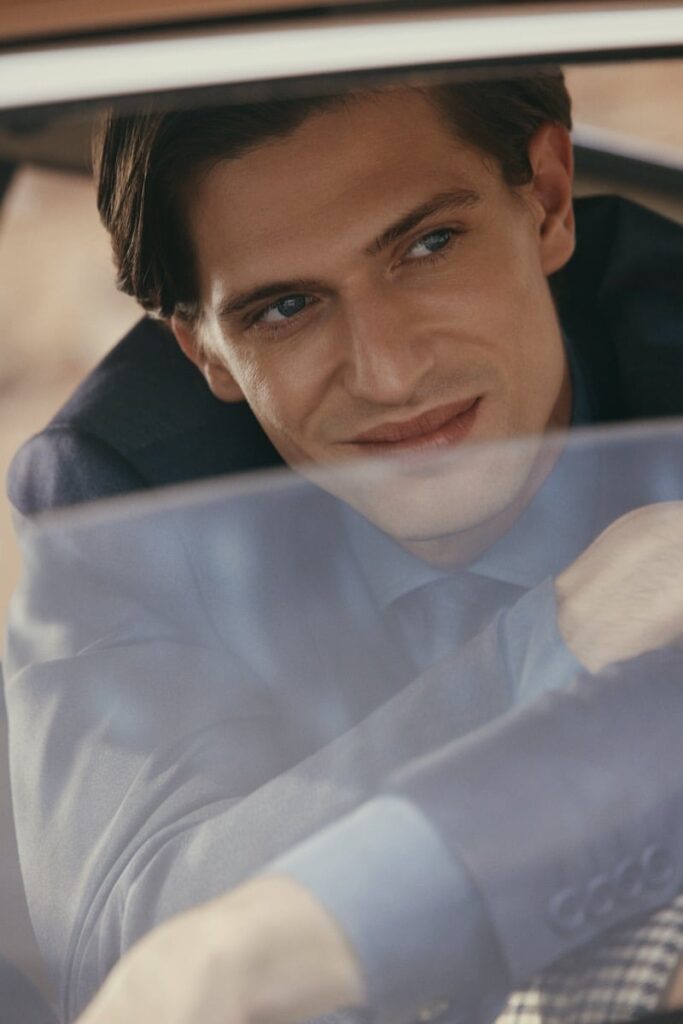 Close-up of a man in a suit looking out through a car window with a slight smile on his face.