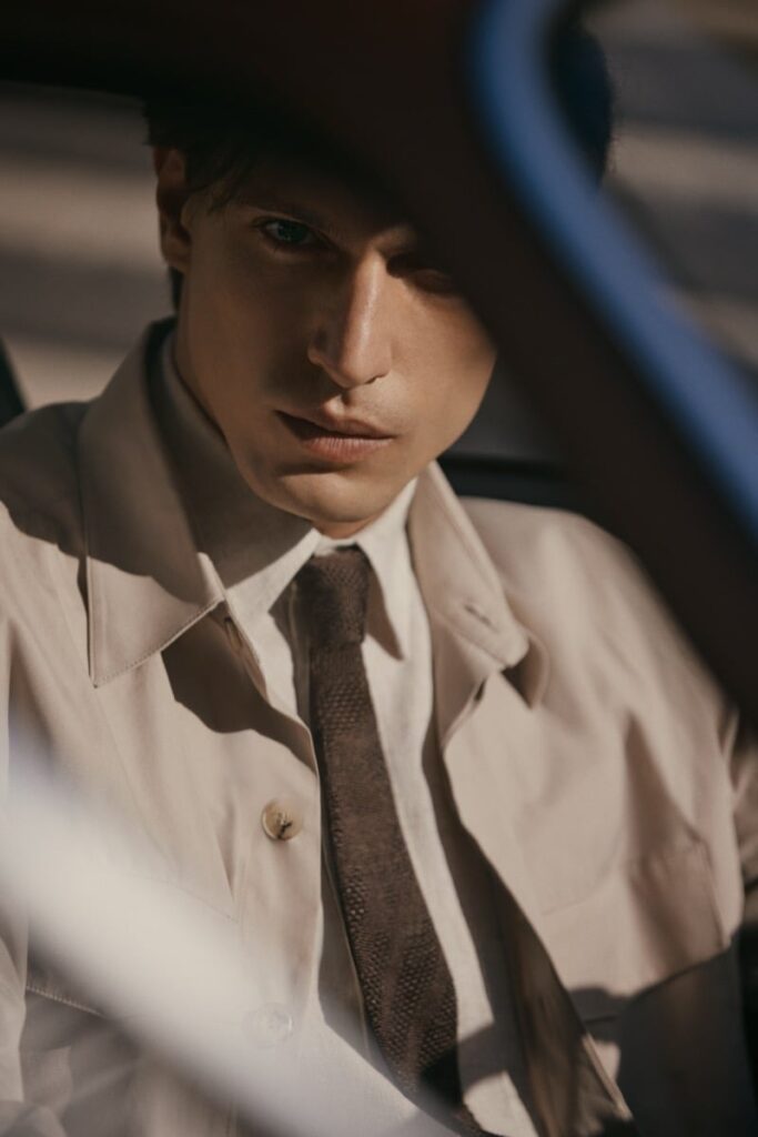 Young man in a beige trench coat and brown tie sits in a car, gazing intently at the camera with dramatic lighting on his face.