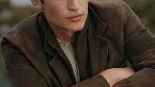 Young man in a brown jacket leaning on a car hood, looking off to the side.