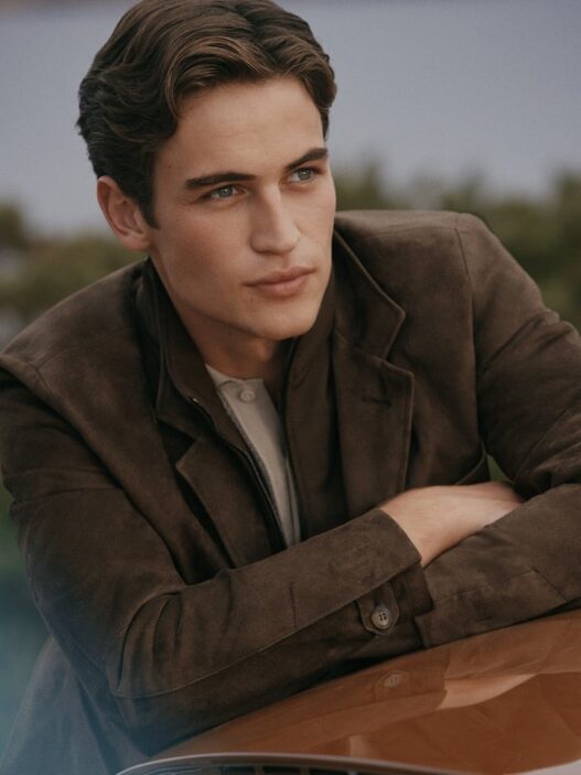 Young man in a brown jacket leaning on a car hood, looking off to the side.
