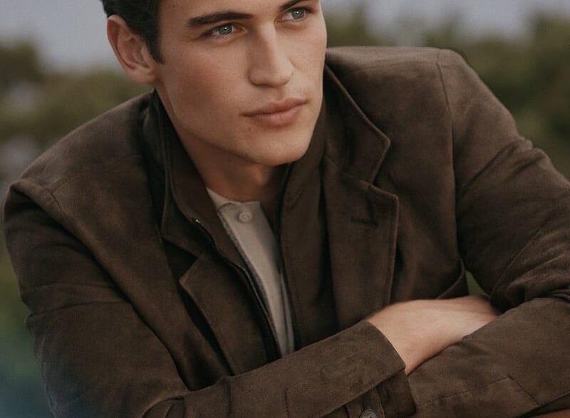 Young man in a brown jacket leaning on a car hood, looking off to the side.