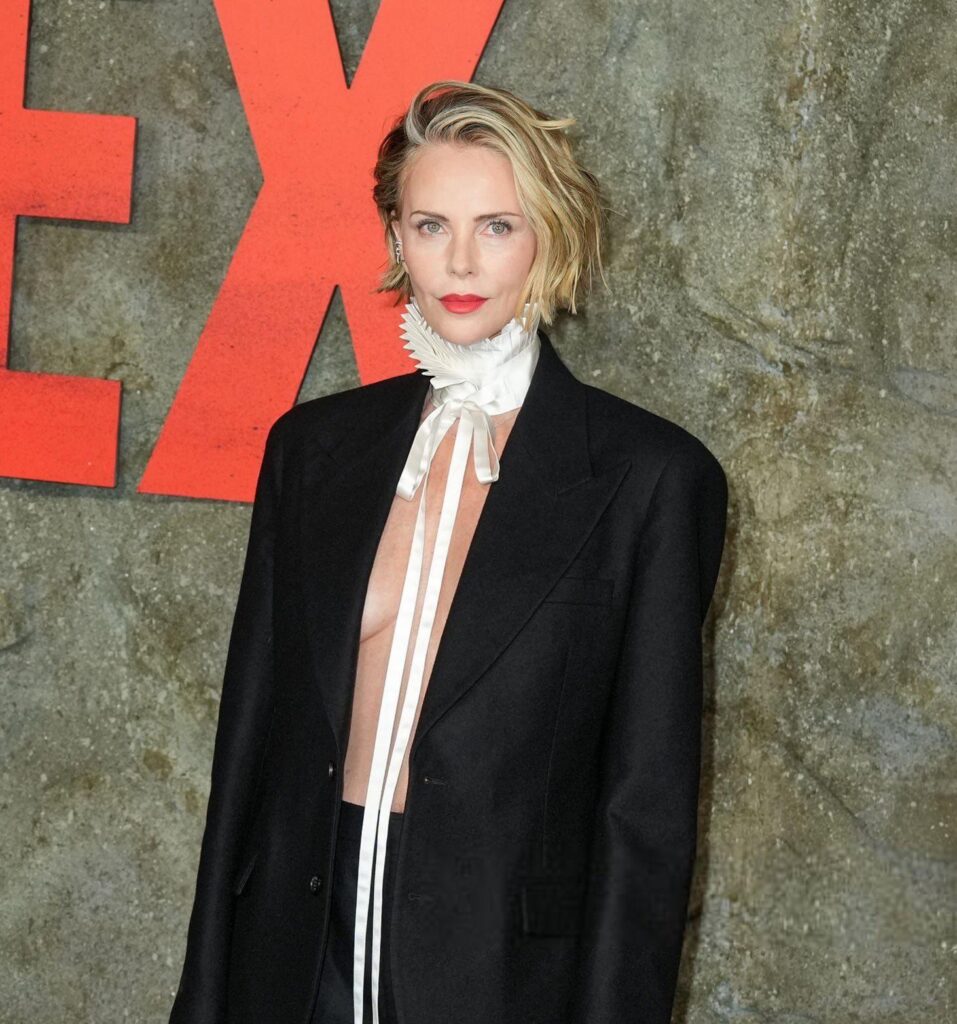 Woman with short blonde hair and red lipstick posing in a black blazer with a white ruffled necktie against a textured wall with large orange letters behind her