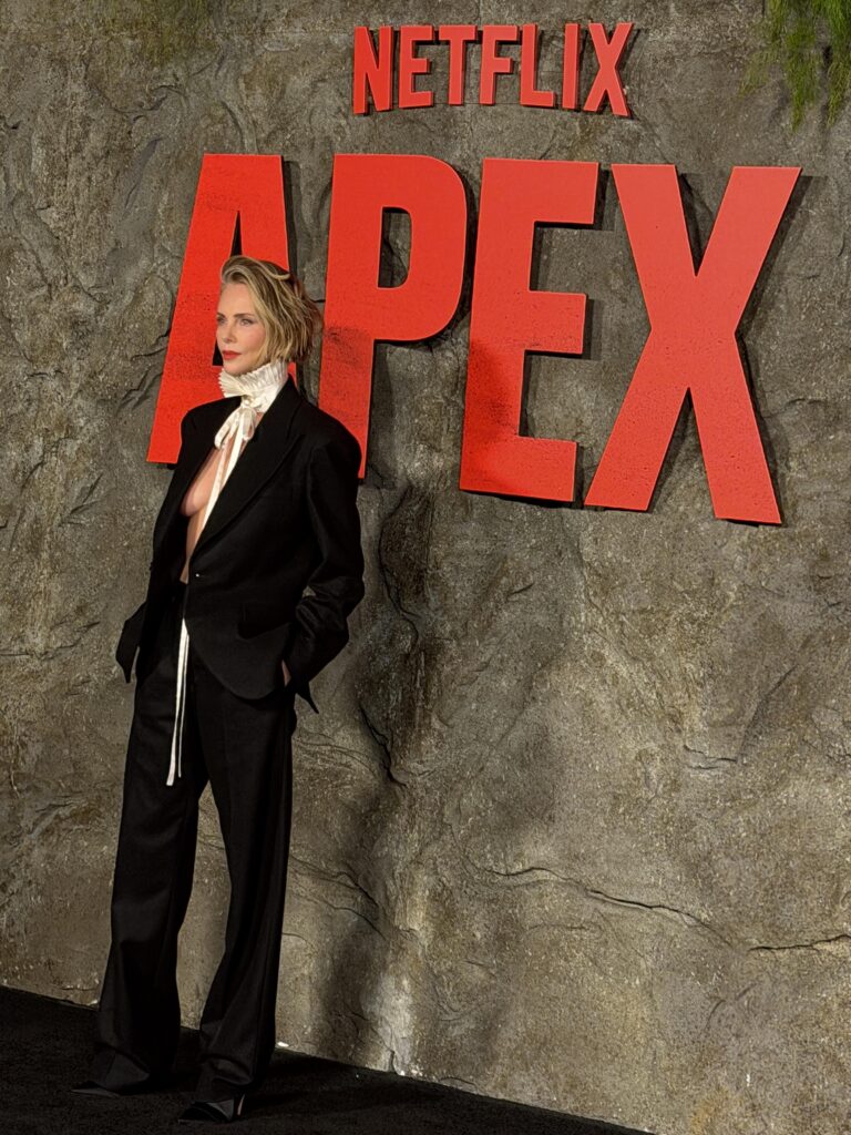 Person in a black suit with a white ruffled scarf posing beside a large red 'APEX' sign and Netflix logo on a rocky wall backdrop.