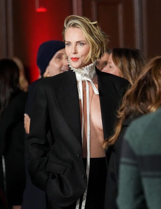 Woman with short blonde hair and red lips wearing a black blazer and white ruffled neck collar at a social event.