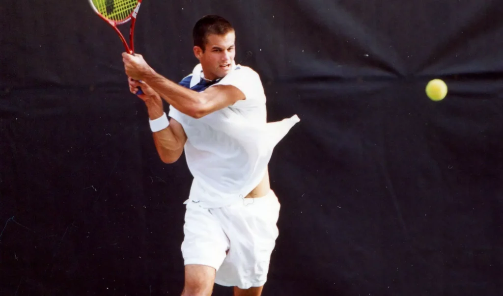 Male tennis player in a white outfit swings a racquet during a backhand stroke as a tennis ball approaches on a dark background.