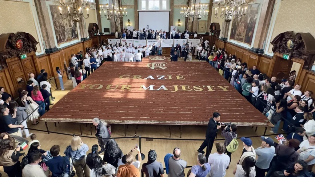 Crowd gathers around a giant wooden stage with the words 'GRAZIE YOUR MAJESTY' printed on it in a grand historic hall.