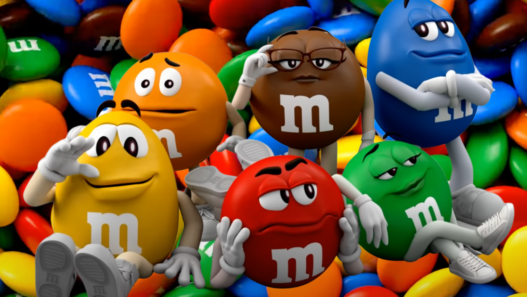 Five anthropomorphic M&M characters posing and lounging on a pile of colorful candies.